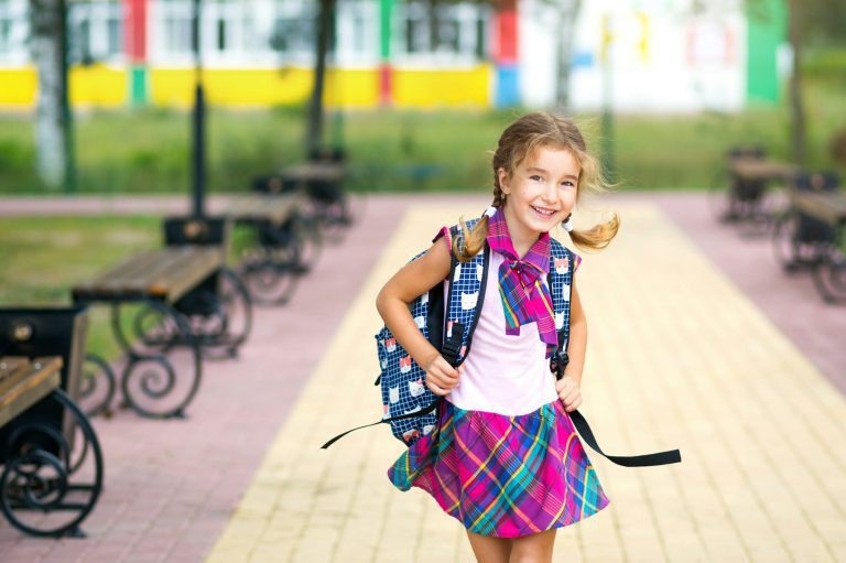 Hogyan segíthet a tanár a mobbing áldozatainak? Cheerful girl with a backpack and in a school uniform in the school yard.
