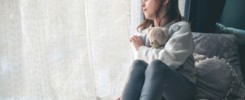 a pensive cute girl sits and looks out the window, hugging a teddy bear.