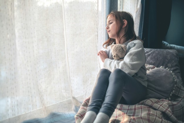 a pensive cute girl sits and looks out the window, hugging a teddy bear.