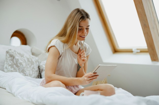 A woman in pajamas is seated on a wooden bed and making notes in note