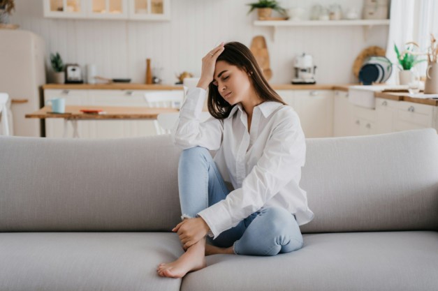 Exhausted brunette hispanic young adult woman in white shirt blue jeans sitting on sofa eyes closed