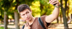 hipster man in brown T-shirt winks and shows class to the phone camera in summer green city park