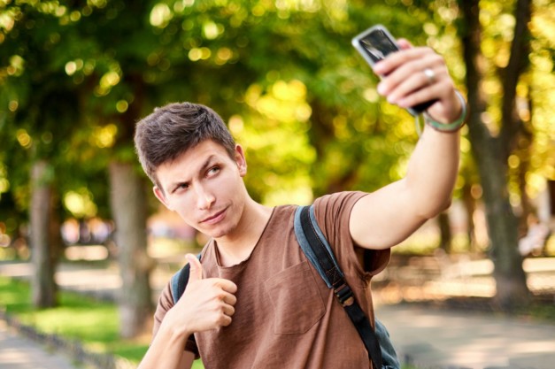 hipster man in brown T-shirt winks and shows class to the phone camera in summer green city park