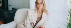 Purposeful blonde woman sitting in chair at home looks at camera dressed in beige suit.