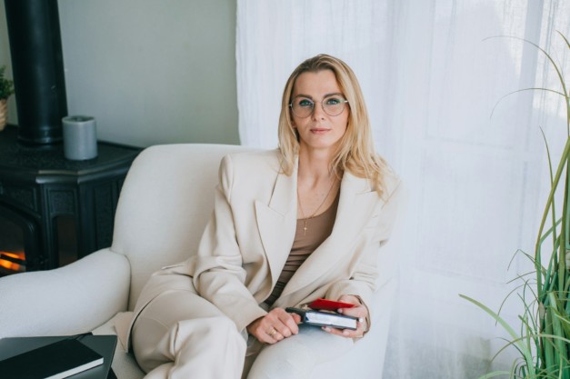 Purposeful blonde woman sitting in chair at home looks at camera dressed in beige suit.