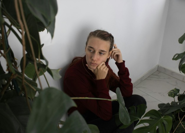 Sad girl in depression sits on the floor in the corner of the room surrounded by plants.