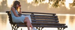 Young sad woman in summer dress sitting alone on lake side bench on warm evening. Solitude and