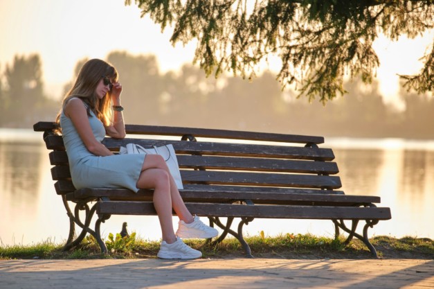 Young sad woman in summer dress sitting alone on lake side bench on warm evening. Solitude and