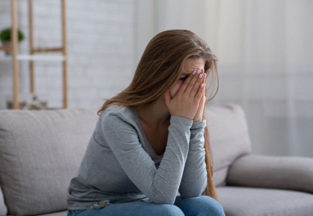 Young woman burying her face in hands and crying, feeling depressed or desperate on sofa indoors